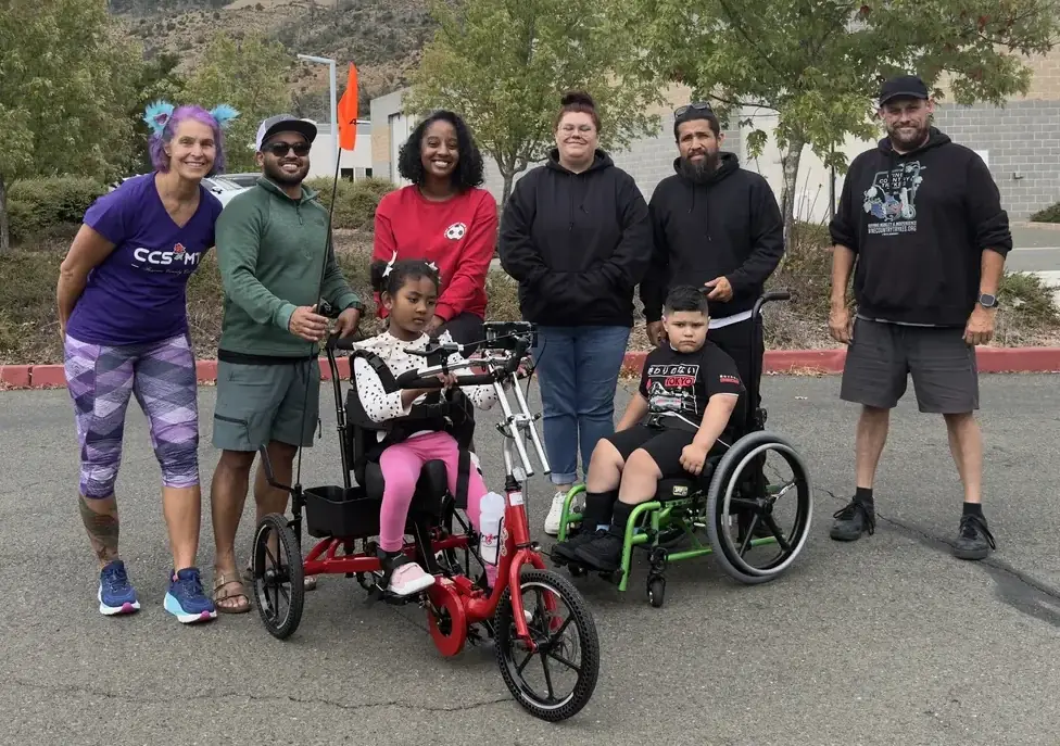 A rider on a red adaptive tricycle with a wheelchair user and community members outdoors.